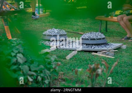 Zwei Metall-Podpeka- oder sache-Deckel mit Holzkohle auf einer Gartenparty. Blick auf typische sache-Töpfe aus Metall, die durch die Blätter eines Gartens blicken. Stockfoto