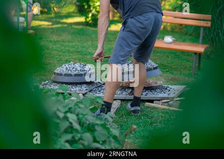 Zwei Metall-Podpeka- oder sache-Deckel mit Holzkohle auf einer Gartenparty. Blick auf typische sache-Töpfe aus Metall, die durch die Blätter eines Gartens blicken. Person li Stockfoto