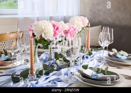 Wunderschöner Tisch. Teller in der Nähe von Gläsern, Pfingstrosen, brennenden Kerzen und Besteck auf dem Tisch im Zimmer Stockfoto