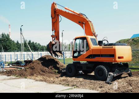 Planierraupenbagger mit Schaufel gräbt Loch. Orangefarbener Traktor mit Eimer und einem Haufen Erde mit Lehm am Sommertag draußen. Stockfoto