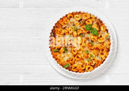 Tortellini gebacken mit Mozzarella-Käse, Tomatensoße und Rinderhack in weißer Backform auf weißem Holztisch, horizontale Sicht von oben, flach liegend Stockfoto