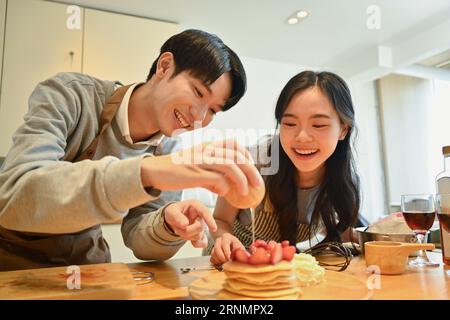 Ein fröhliches junges Paar, das Honig auf leckere Pfannkuchen mit Erdbeeren auf der hölzernen Küchenplatte gießt Stockfoto