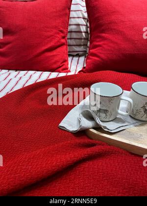 Rote Bettwäsche. Holztablett mit Keramikbechern auf dem Bett. Frühstück im Bett. Natürliche Materialien aus Baumwolle und Leinen. Die Romantik der Liebe. Weiße Tassen Stockfoto