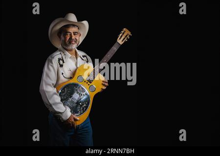 Das Porträt des bärtigen asiatischen Musikers trägt Cowboy-Kleidung, hält an einer Dobro-Gitarre fest und vereint Americana- und Eastern-Stile auf schwarzem Hintergrund Stockfoto