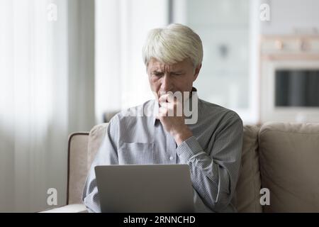 Die Stirn runzelnd besorgt sich der alte Mann, der zu Hause einen Laptop benutzt Stockfoto