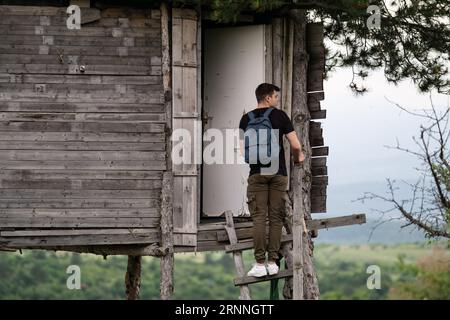 Hübscher junger Mann mit Rucksack, der auf der Treppe eines wilden Baumhauses steht und Fotos mit seinem Handy macht. Wanderer vor dem Baumhaus Stockfoto