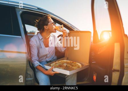 Eine Frau mittleren Alters isst gerade italienische Pizza, sitzt auf dem Fahrersitz während der Essenspause und genießt den Sonnenuntergang. Autofahrt, Fast Food je Stockfoto