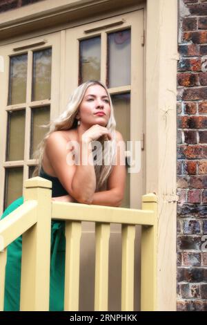 Blonde Frau mit langen Haaren und grünem Outfit posiert vor einem Haus Stockfoto