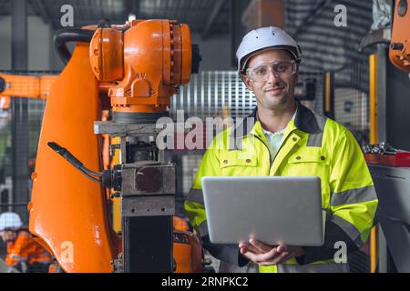 Porträt glücklicher Ingenieur männlich mit Laptop-Computer-Steuerung bedienen Industrie-Roboter Arm Maschine. Man Programmierservice Schweißroboter in Auto Stockfoto