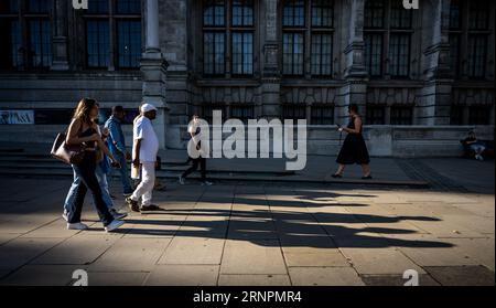 London, Großbritannien: Menschen, die auf dem Bürgersteig spazieren gehen und die Abendsonne lange Schatten wirft. Vor dem Victoria and Albert Museum in South Kensington. Stockfoto