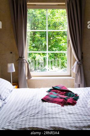 Teilweiser Blick auf ein Zimmer. Ein Bett mit weißen Laken und einer schottischen Schottendecke auf der Oberseite, Tischleuchte auf einer Seite. Im Hintergrund ein weiß gerahmtes Fenster Stockfoto