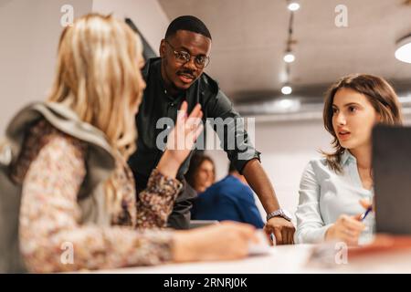 Teamleiterin mittleren Alters, blond, erklärt Projektanweisungen und Arbeitsabläufe ihren verschiedenen jüngeren Kollegen. Schulung bei der Arbeit. Ausbildung am Arbeitsplatz. Cla Stockfoto