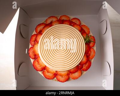 Veganer Geburtstagskuchen mit Vanille und Erdbeeren bedeckt mit weißer Schokolade in einem geöffneten Karton auf einem Tisch und weißem Hintergrund Stockfoto