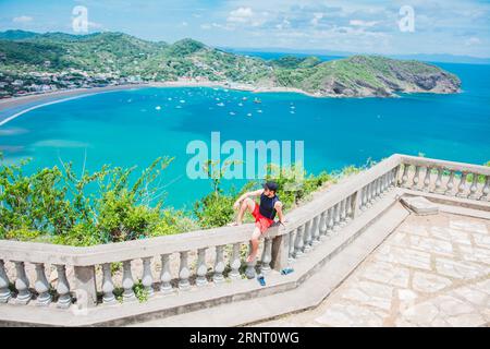 Glücklicher Tourist, der in einem Aussichtspunkt auf eine Bucht sitzt. Porträt des Touristen-Mannes im Blick auf die Bucht von San Juan del Sur. Reise- und Tourismusförderung Stockfoto