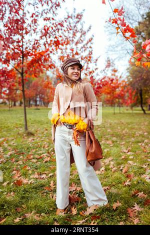 Ganzkörperporträt einer jungen Frau, die im Herbstpark unter roten Bäumen einen stylischen warmen Pulli mit Hut und Handtasche trägt. Modisches Damen-Outfit. Stockfoto