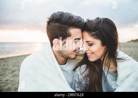 Ein liebevolles Paar, das am Strand sitzt und von einer weißen Decke bedeckt ist Stockfoto