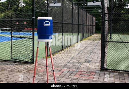 Blaue Wasserkanne auf einem Stativ neben den Tennis- und Picklball-Plätzen, umgeben von schwarzen Zäunen. Stockfoto