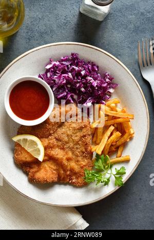 Schnitzel mit Kartoffelfrites, Rotkohlsalat und Sauce auf weißem Teller auf dunklem Steinhintergrund. Draufsicht, flach Stockfoto