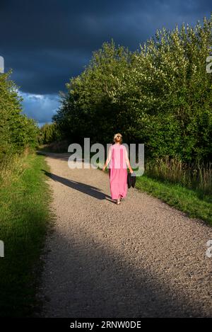 Blonde Frau in einem rosa Kleid, die auf dem Land mit abendlichen hellen, langen Schatten spaziert Stockfoto