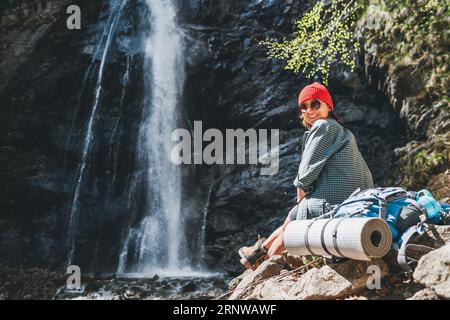Lächelnde Frau mit Rucksack in rotem Hut in aktiver Trekkingkleidung und Stiefeln, die in der Nähe des Gebirgswasserfalls sitzt und die Natur genießt Stockfoto
