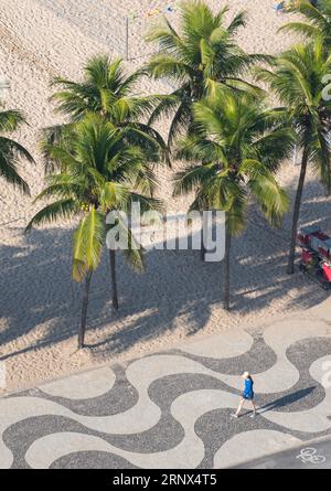 Rio de Janeiro, Brasilien: Eine Frau, die auf dem ikonischen grafischen Bürgersteig des Copacabana Strandes entlang der Avenida Atlantica (Atlantic Avenue), Sand und Palmen spaziert Stockfoto