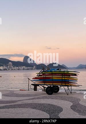 Rio de Janeiro, Brasilien: Surfbretter stapeln sich auf einem Wagen bei Sonnenuntergang am Strand von Ipanema mit dem ikonischen grafischen Bürgersteig entlang der Avenida Atlantica Stockfoto