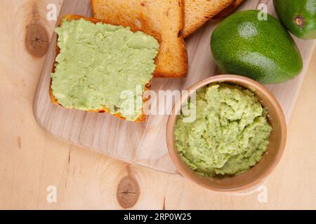 Auf der blassgelben Holzoberfläche finden Sie Stillleben mit Avocadotoast, üppigem Avocadoaufstrich und zwei perfekt frischen Avocadofrüchten aus nächster Nähe Stockfoto