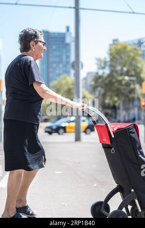 Eine ältere Frau, die im Sommer die Straße mit dem Einkaufswagen überquert Stockfoto