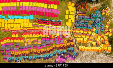 Narayanganj, Dhaka, Bangladesch. September 2023. Hunderte von gefärbten Stoffstücken verteilen sich auf einem Feld im Batik Village in Narayanganj, Bangladesch zum Trocknen, das wie ein Königreich der Farben aussieht. Arbeiter verwenden Hüte zum Schutz vor der sengenden Hitze, da sie die bunten Stoffe ständig drehen müssen, damit sie im Sonnenlicht perfekt trocknen. Wunderschön verzierte bunte Tücher werden mit der indonesischen Technik namens „Batik“ hergestellt. Teile der Konstruktion werden durch Auftragen von heißem Wachs blockiert, dann wird ein Farbstoff darauf aufgetragen und die mit Wachs bedeckten Teile sind widerstandsfähig Stockfoto