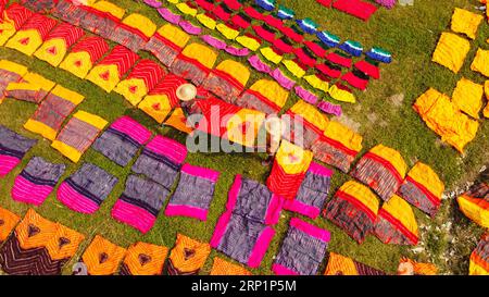 Narayanganj, Dhaka, Bangladesch. September 2023. Hunderte von gefärbten Stoffstücken verteilen sich auf einem Feld im Batik Village in Narayanganj, Bangladesch zum Trocknen, das wie ein Königreich der Farben aussieht. Arbeiter verwenden Hüte zum Schutz vor der sengenden Hitze, da sie die bunten Stoffe ständig drehen müssen, damit sie im Sonnenlicht perfekt trocknen. Wunderschön verzierte bunte Tücher werden mit der indonesischen Technik namens „Batik“ hergestellt. Teile der Konstruktion werden durch Auftragen von heißem Wachs blockiert, dann wird ein Farbstoff darauf aufgetragen und die mit Wachs bedeckten Teile sind widerstandsfähig Stockfoto