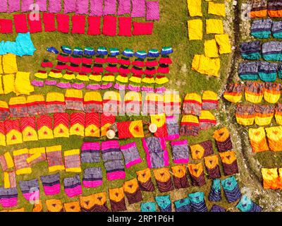 Narayanganj, Dhaka, Bangladesch. September 2023. Hunderte von gefärbten Stoffstücken verteilen sich auf einem Feld im Batik Village in Narayanganj, Bangladesch zum Trocknen, das wie ein Königreich der Farben aussieht. Arbeiter verwenden Hüte zum Schutz vor der sengenden Hitze, da sie die bunten Stoffe ständig drehen müssen, damit sie im Sonnenlicht perfekt trocknen. Wunderschön verzierte bunte Tücher werden mit der indonesischen Technik namens „Batik“ hergestellt. Teile der Konstruktion werden durch Auftragen von heißem Wachs blockiert, dann wird ein Farbstoff darauf aufgetragen und die mit Wachs bedeckten Teile sind widerstandsfähig Stockfoto