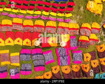 Narayanganj, Dhaka, Bangladesch. September 2023. Hunderte von gefärbten Stoffstücken verteilen sich auf einem Feld im Batik Village in Narayanganj, Bangladesch zum Trocknen, das wie ein Königreich der Farben aussieht. Arbeiter verwenden Hüte zum Schutz vor der sengenden Hitze, da sie die bunten Stoffe ständig drehen müssen, damit sie im Sonnenlicht perfekt trocknen. Wunderschön verzierte bunte Tücher werden mit der indonesischen Technik namens „Batik“ hergestellt. Teile der Konstruktion werden durch Auftragen von heißem Wachs blockiert, dann wird ein Farbstoff darauf aufgetragen und die mit Wachs bedeckten Teile sind widerstandsfähig Stockfoto