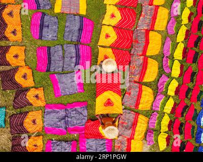 Narayanganj, Dhaka, Bangladesch. September 2023. Hunderte von gefärbten Stoffstücken verteilen sich auf einem Feld im Batik Village in Narayanganj, Bangladesch zum Trocknen, das wie ein Königreich der Farben aussieht. Arbeiter verwenden Hüte zum Schutz vor der sengenden Hitze, da sie die bunten Stoffe ständig drehen müssen, damit sie im Sonnenlicht perfekt trocknen. Wunderschön verzierte bunte Tücher werden mit der indonesischen Technik namens „Batik“ hergestellt. Teile der Konstruktion werden durch Auftragen von heißem Wachs blockiert, dann wird ein Farbstoff darauf aufgetragen und die mit Wachs bedeckten Teile sind widerstandsfähig Stockfoto