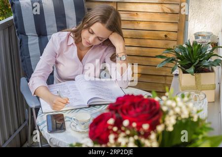 Attraktive junge Frau, die hart zu den Prüfungen lernt. Lernkonzept. Rote Rosen im Vordergrund. Auf dem Balkon Stockfoto