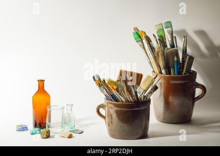 Künstlerische Pinsel, bunte schöne Steine, alte Flaschen und Malwerkzeuge in alten Keramiktöpfen auf weißem Hintergrund. Künstlerische Komposition Stockfoto