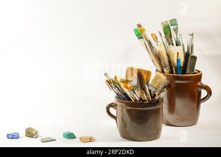 Künstlerische Pinsel, bunte schöne Steine und Malwerkzeuge in alten Keramiktöpfen auf weißem Hintergrund. Künstlerische Komposition Stockfoto