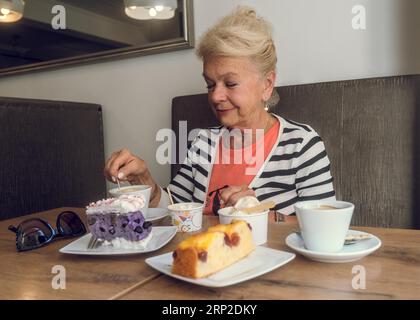 Attraktive, freudige ältere Frau, die Kaffee trinkt, während sie in einem Café sitzt. Porträt einer Reifen Frau in einer Cafeteria. Bunte Kuchen und Kaffee auf dem Stockfoto