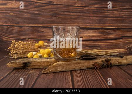 Ein wunderschönes geschliffenes Glas, gefüllt mit einer bernsteinfarbenen Flüssigkeit auf einem Holzstück neben einigen Anissternen, getrockneten Blumen und viel Holz Stockfoto