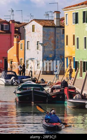 Insel Burano in der Lagune von Venedig, Gemeinde Venedig, Italien. Zwei Männer paddeln mit dem Schlauchboot den Kanal hinunter. Farbenfrohe Häuser. Stockfoto