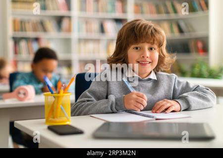 Positiver, intelligenter Schuljunge, der im Klassenzimmer am Schreibtisch sitzt und in der Kamera lächelt, während er im Notebook schreibt Stockfoto