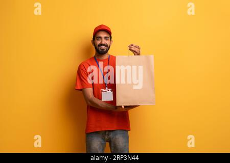 Glücklicher indischer Liefermann in Uniform, der die Handwerkspapiertasche vor der Kamera zeigt Stockfoto