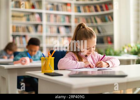 Gruppe von intelligenten Schulkindern, die an Schreibtischen im Klassenzimmer oder in der Bibliothek in der Grundschule sitzen und in ihrem Arbeitsbuch schreiben Stockfoto