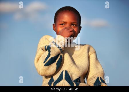 Außenporträt eines glücklichen Kindes in einem afrikanischen Dorf, das seine Nase hält, tagsüber sonniger Tag mit flachem Bokeh Stockfoto