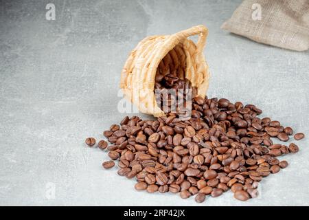 Ein Haufen Kaffeebohnen, der aus einem Korb aus Zuckerrohr auf einem grauen abstrakten Hintergrund auslief. Stockfoto