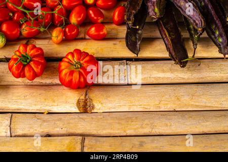Verschiedene rote Tomaten und dunkel-violette geflügelte Bohnenschoten auf einer rustikalen Tischplatte aus Bambusholz mit Kopierraum Stockfoto