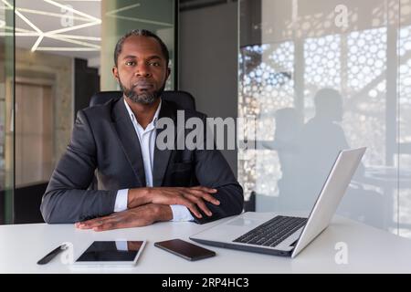 Porträt von ernsthaftem Denken und konzentriertem Boss, afroamerikanischer Mann, der die Kamera mit gefalteten Händen auf dem Schreibtisch betrachtet, Boss, der mit Laptop im Büro arbeitet, Investor, der sich des Sieges sicher ist. Stockfoto