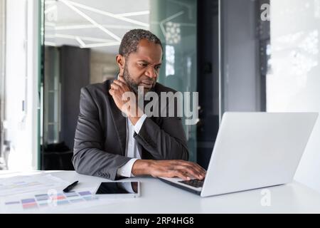 Ausgereifter Geschäftsmann mit leichtem Denken am Arbeitsplatz im Büro, afroamerikanischer grauhaariger Boss, der mit Laptop im Büro arbeitet. Stockfoto