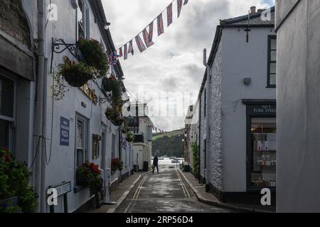 Forestreet Salcombe an einem bewölkten Sommertag mit Union Jack Fahnenfliegen, während eine Einzelperson mit Fortescue Pub zwischen Pfützen auf der Straße spaziert Stockfoto