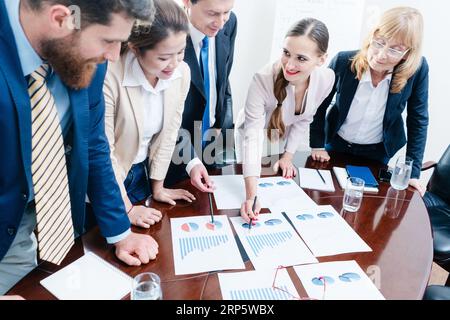 Ein multiethnisches Team aus fünf engagierten Spezialisten lächelt, während es verschiedene Diagramme mit positiven Statistiken analysiert Stockfoto