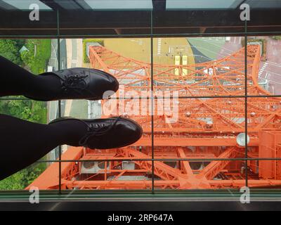 Weibliche Füße mit schwarzen Schuhen und Strumpfhosen stehen auf einem Glasboden im Tokio Tower in Japan. Blick auf das vom Eiffelturm inspirierte Observatorium. Stockfoto